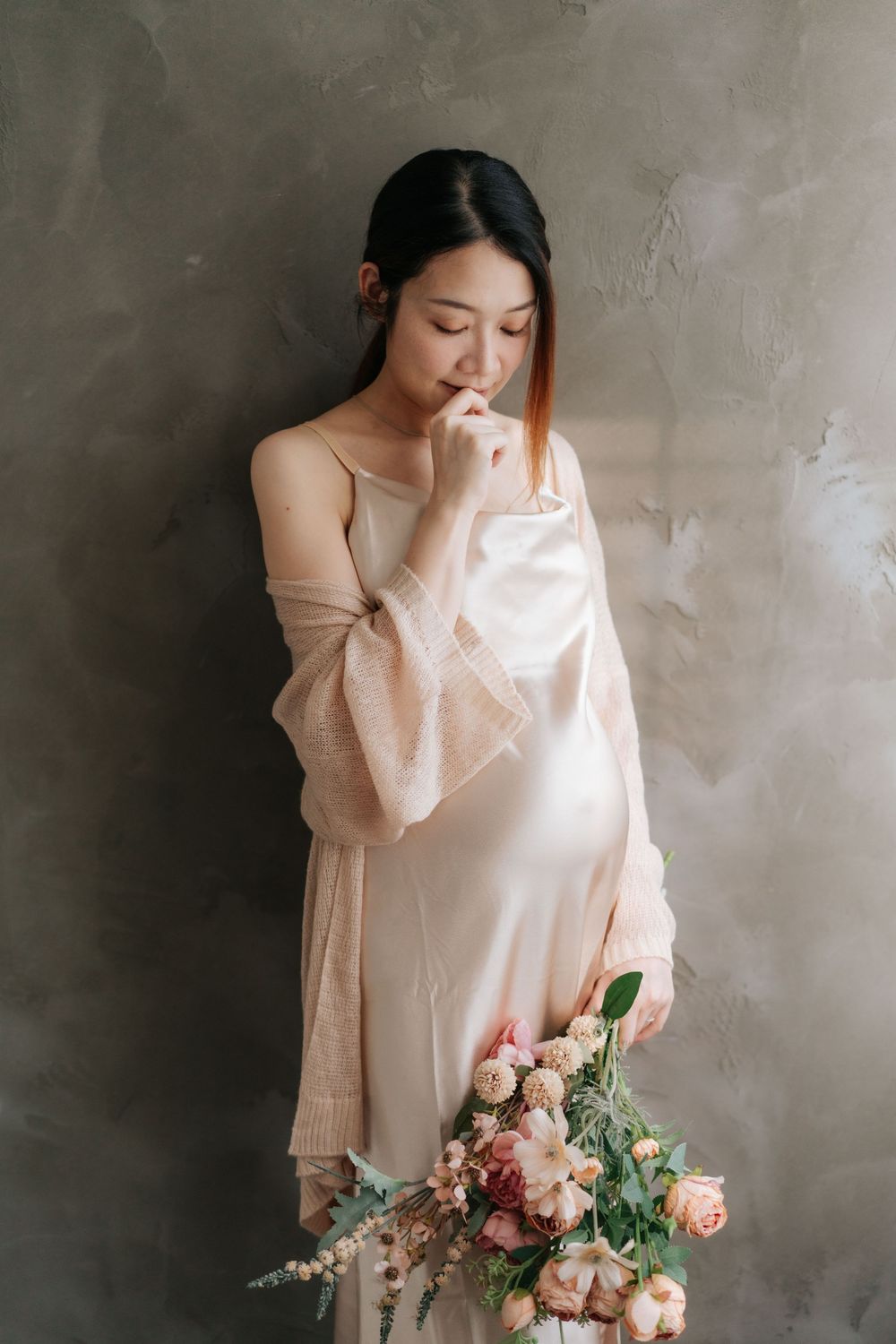 A person in a light pink dress and cardigan stands thoughtfully against a textured wall while holding a bouquet of pink flowers.