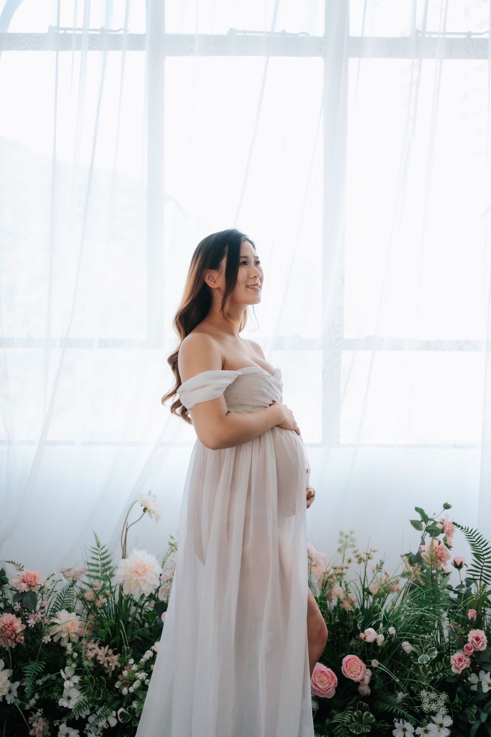 A maternity photoshoot featuring soft pink roses and white curtains creating an ethereal indoor garden setting.