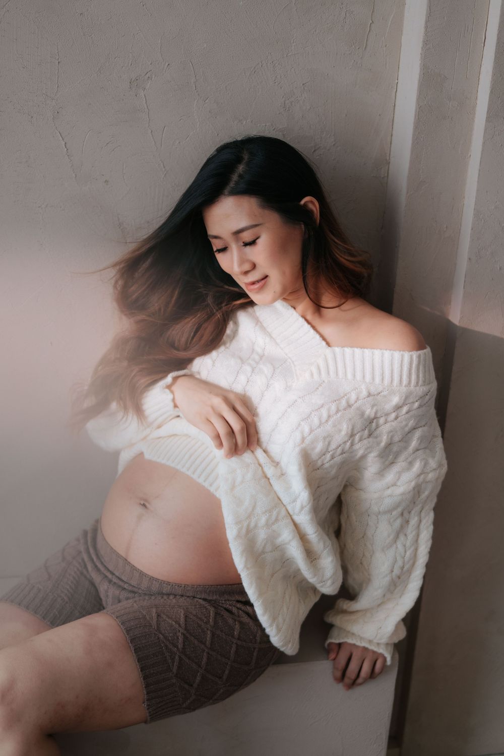 A series of maternity photos showing a pregnant woman in a white sweater lying against a gray wall.