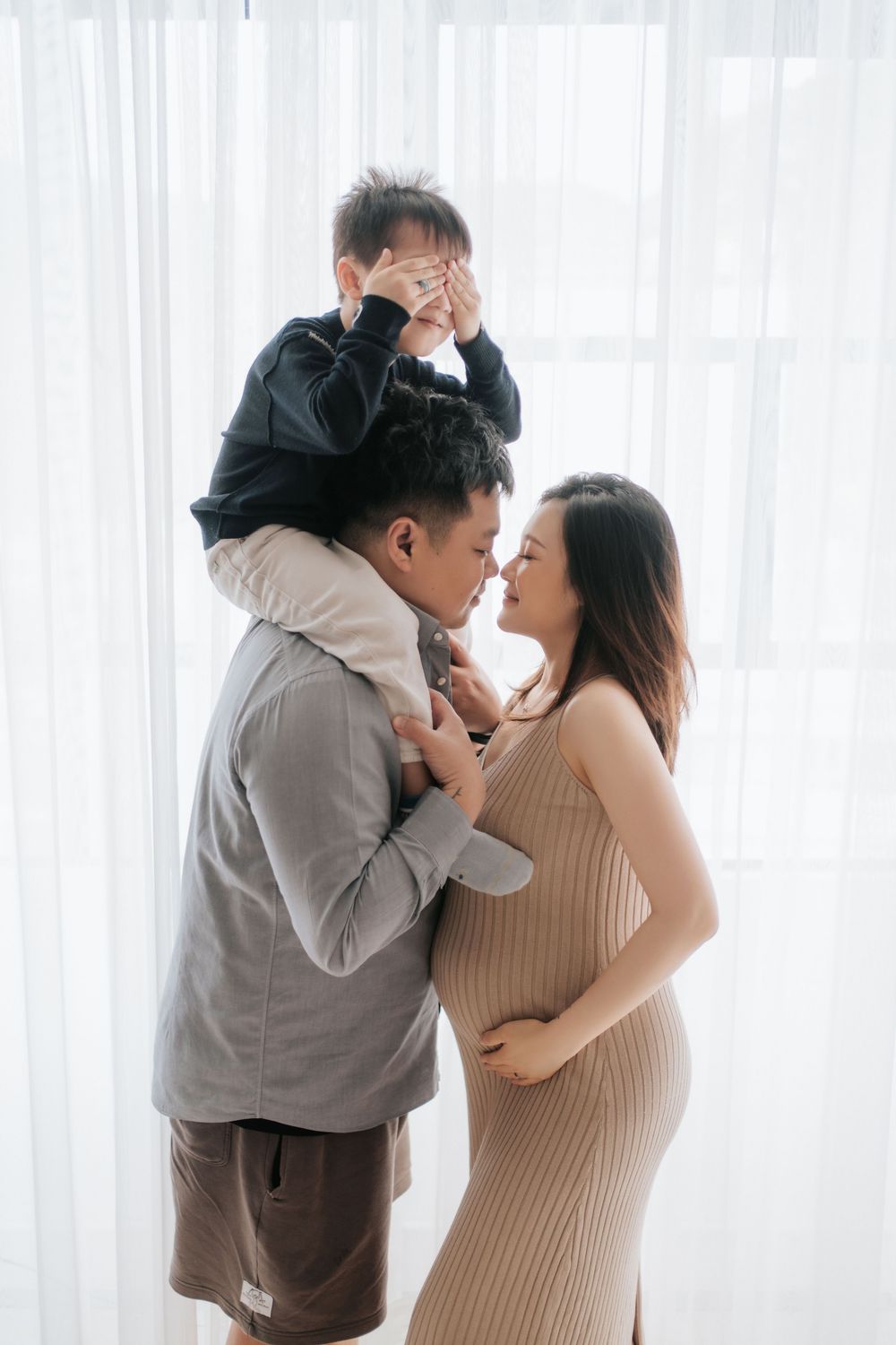 A family shares tender moments in a bright room with white curtains, wearing grey and beige outfits.