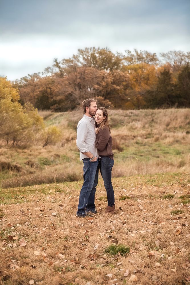 A couple shares a romantic moment in a rustic autumn field surrounded by golden foliage and tall trees.