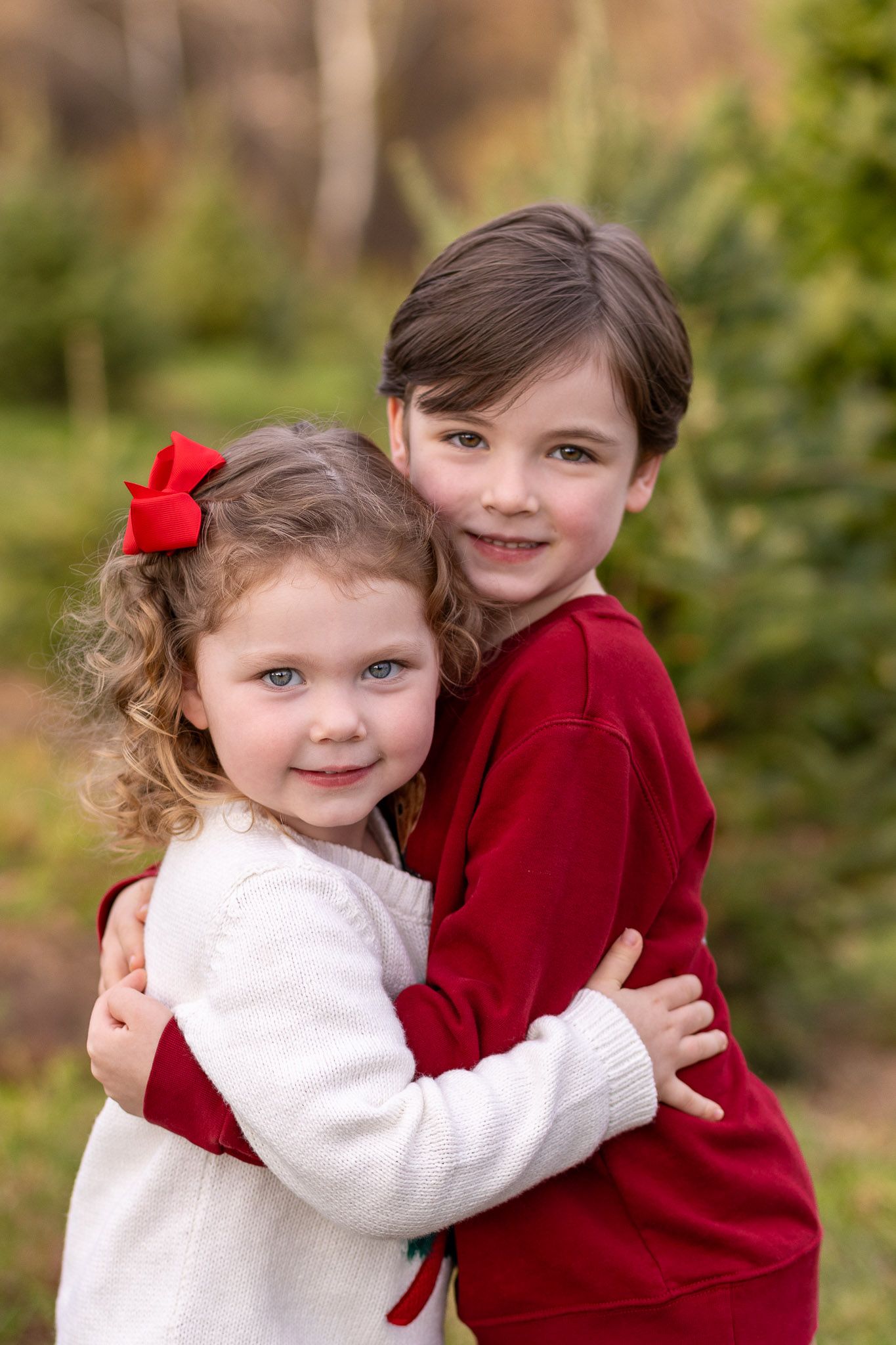 Two children in red and white outfits share a sweet hug while posing outdoors in a natural setting.