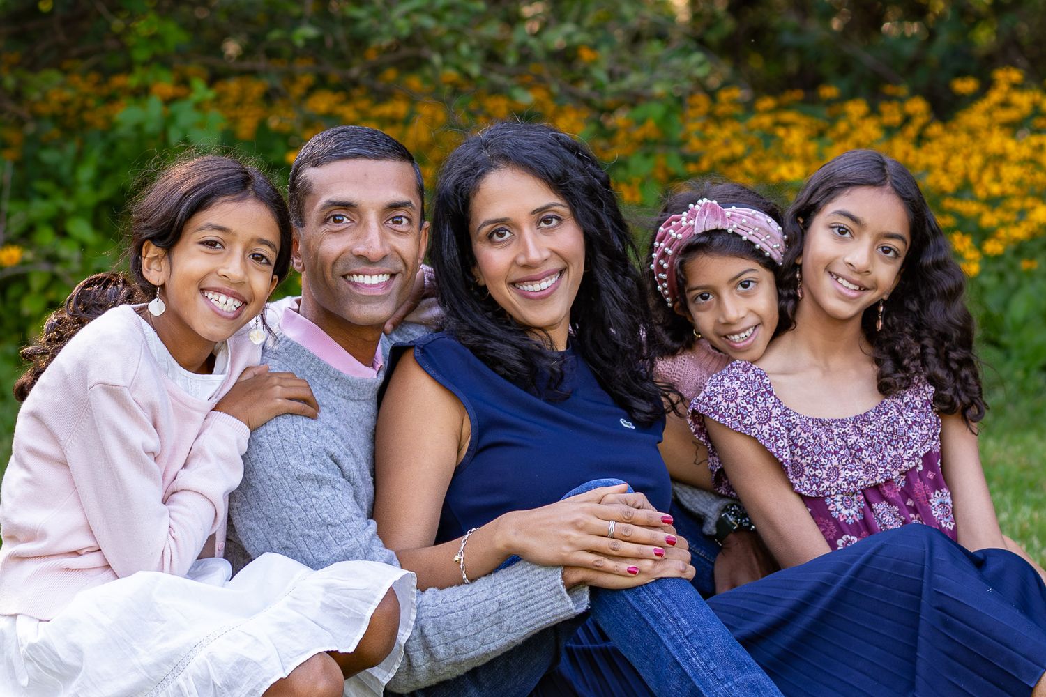 A family sits together outdoors against a backdrop of vibrant yellow autumn flowers in a garden setting.