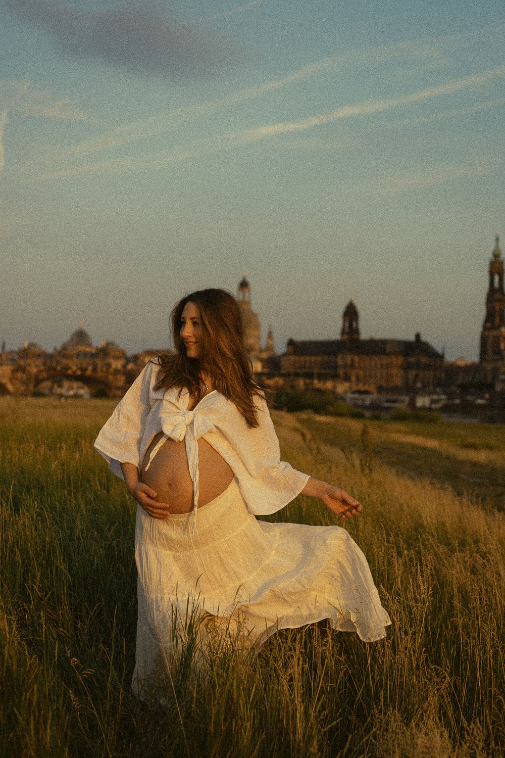 Portrait Fotoshooting Dresden - Frau präsentiert nackten Babybauch auf Elbiesen bei Sonnenuntergang mit Altadt von Dresden im Hintergrund