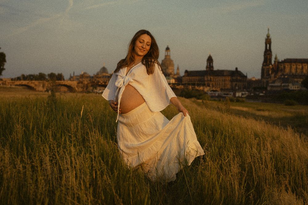 Schwangere Frau hält sich Rock an Saum fest und läuft über die Elbwiesen zur Goldenen Stunde - Babybauchfotoshooting Altstadt Dresden