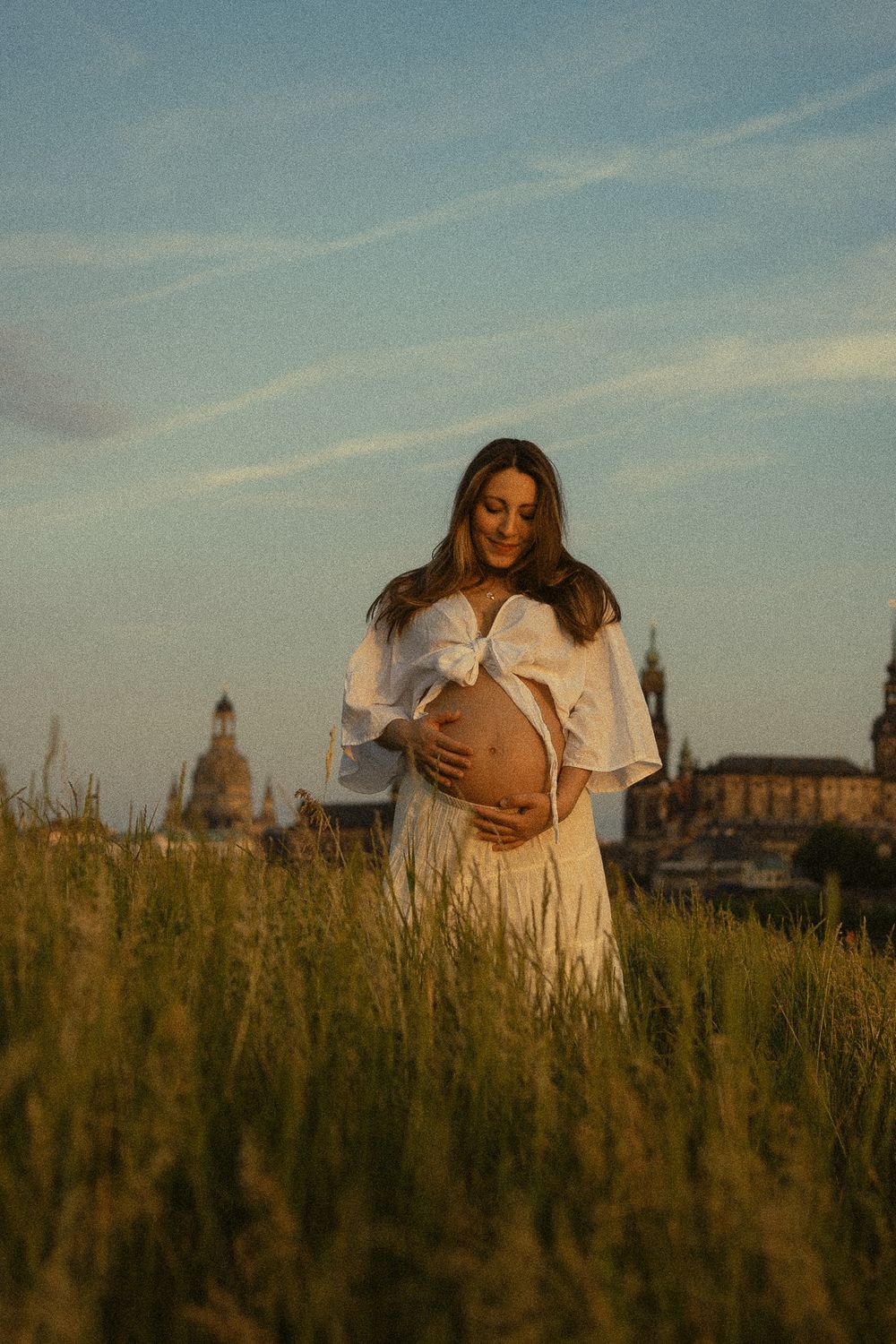 Frau fässt mit beiden Händen an den Babybauch und guckt lächelnd hinunter - Vintage Babybauchfotoshooting Elbwiesen Dresden