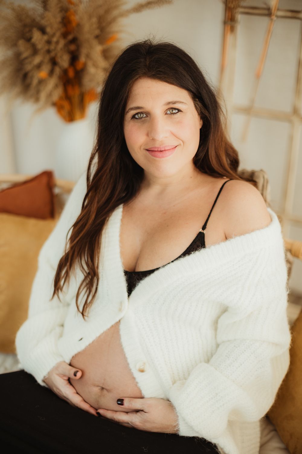 A maternity photo series showing a person in a white sweater cradling their pregnant belly in warm lighting.
