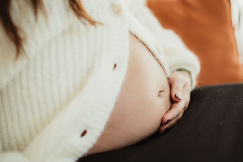 A close-up view of a pregnant woman's bare belly in a white sweater against a warm brown background.