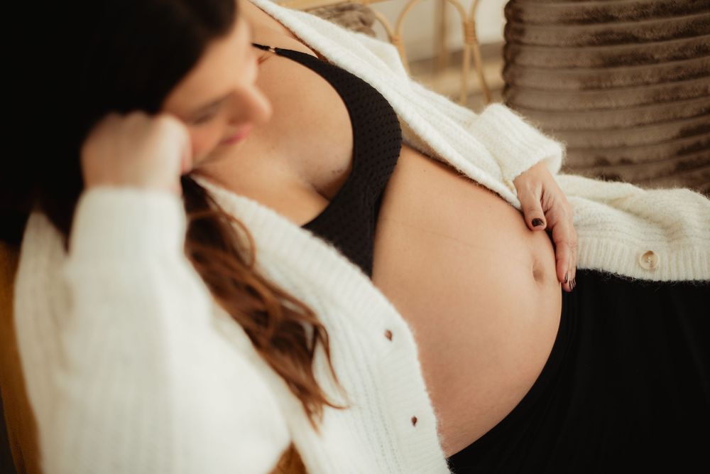 Pregnant woman in white cardigan gently touching her baby bump in soft natural light.