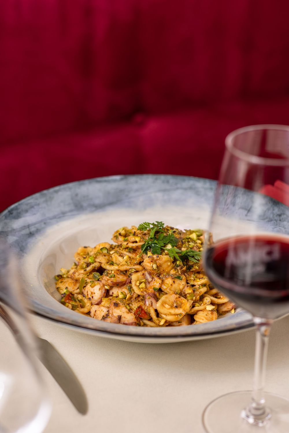 Plated pasta dish with mushrooms and herbs served alongside a glass of red wine on a white table.