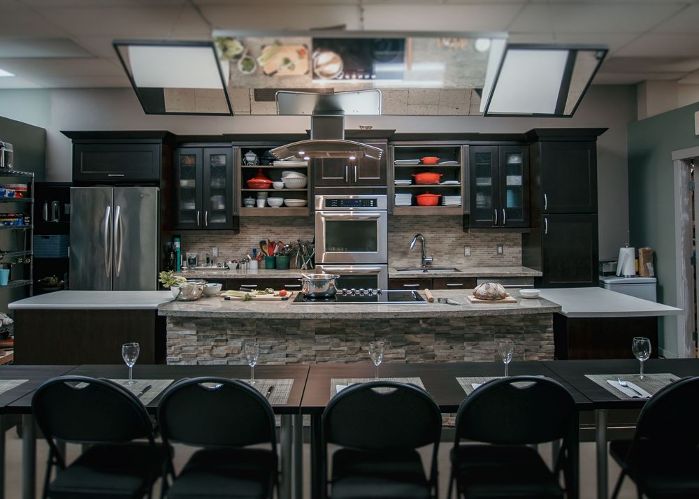 Empty commercial kitchen with stainless steel prep tables and black chairs in a dark room with overhead lighting.