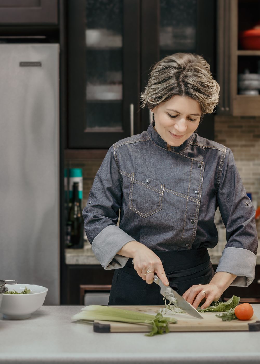 Chef in denim jacket and black apron carefully chopping fresh celery and vegetables in modern kitchen.