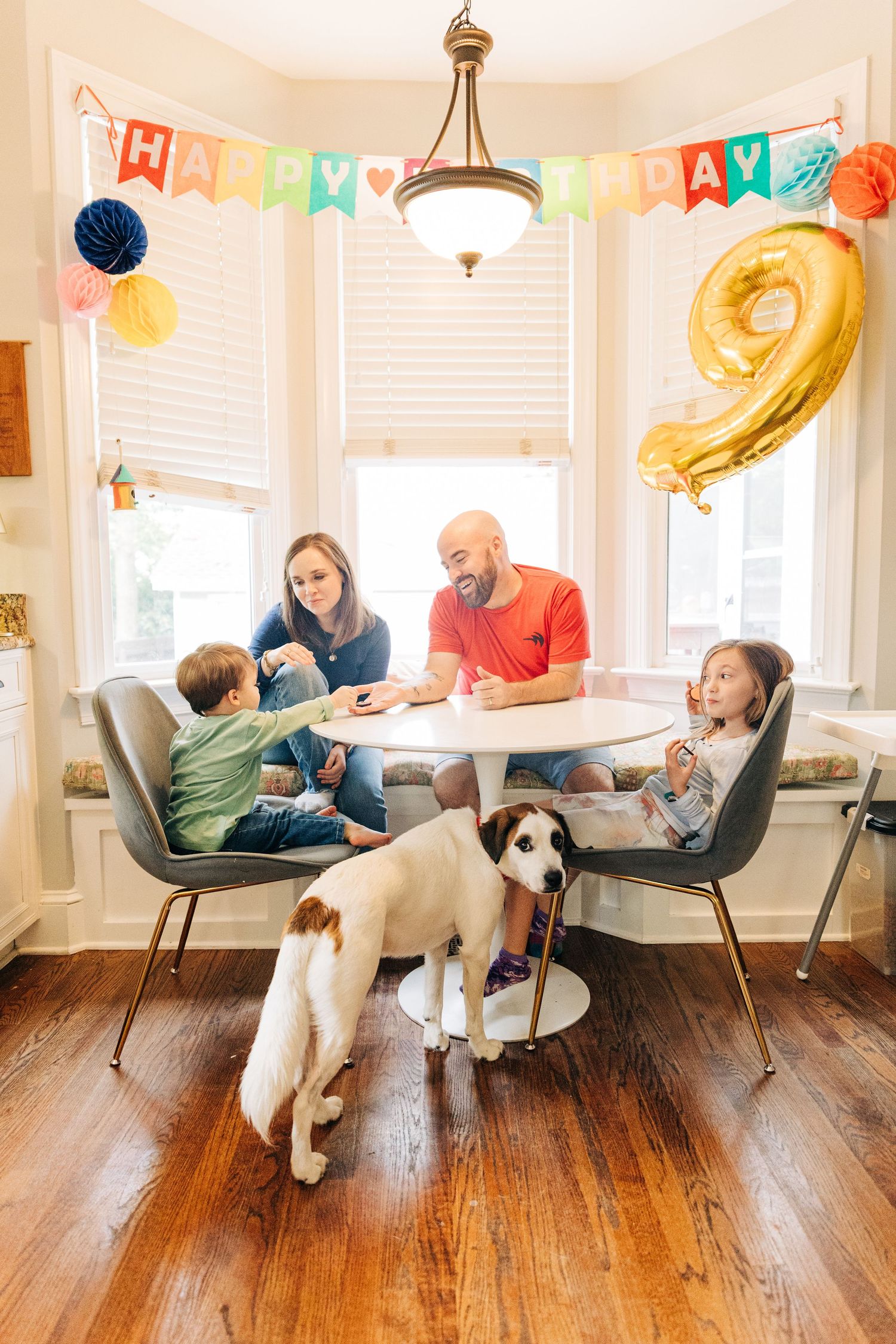 Family gathered around table for birthday celebration with colorful banner and gold number 9 balloon while dog watches below.