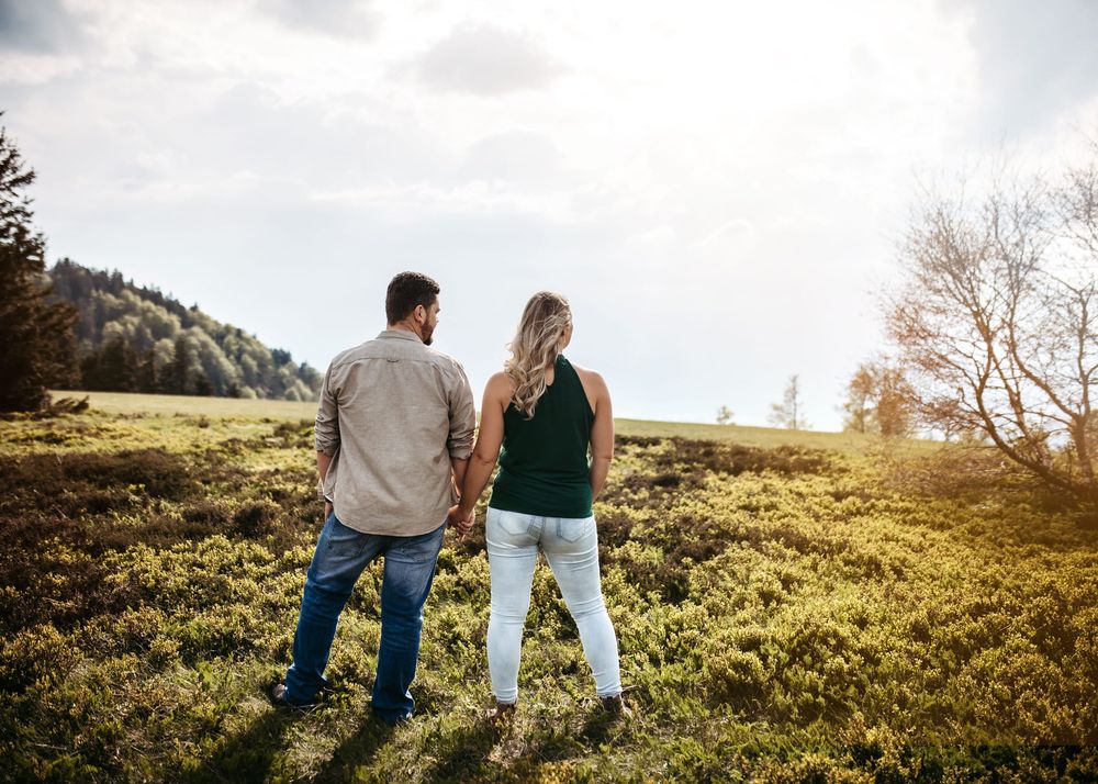 A couple stands together on a grassy hillside overlooking a scenic mountain landscape on a sunny afternoon.