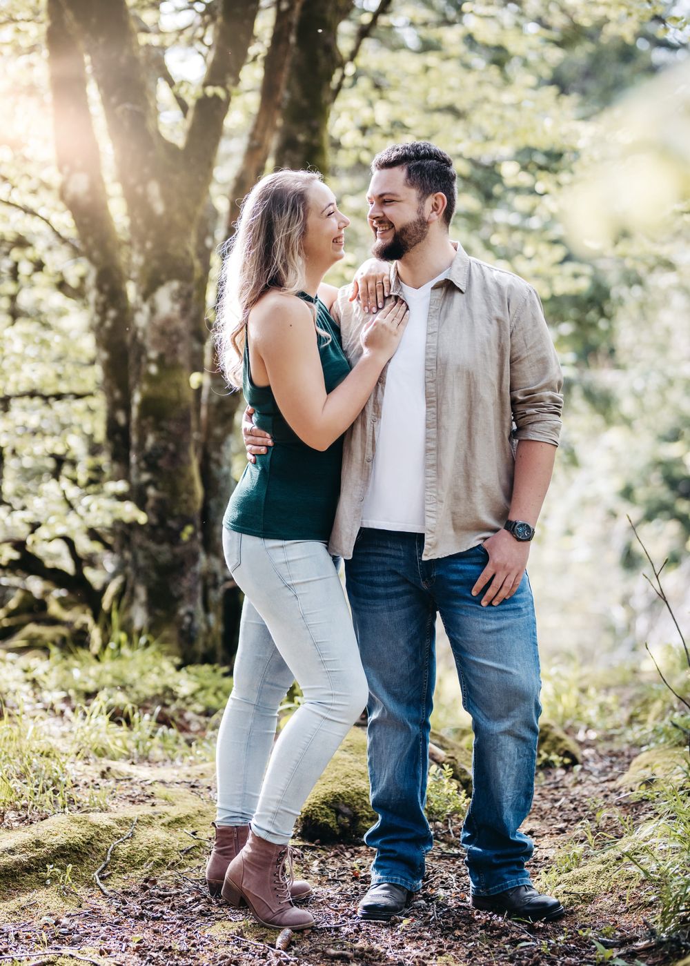 A couple shares a romantic moment in a sunlit forest setting wearing casual clothing and smiling together.