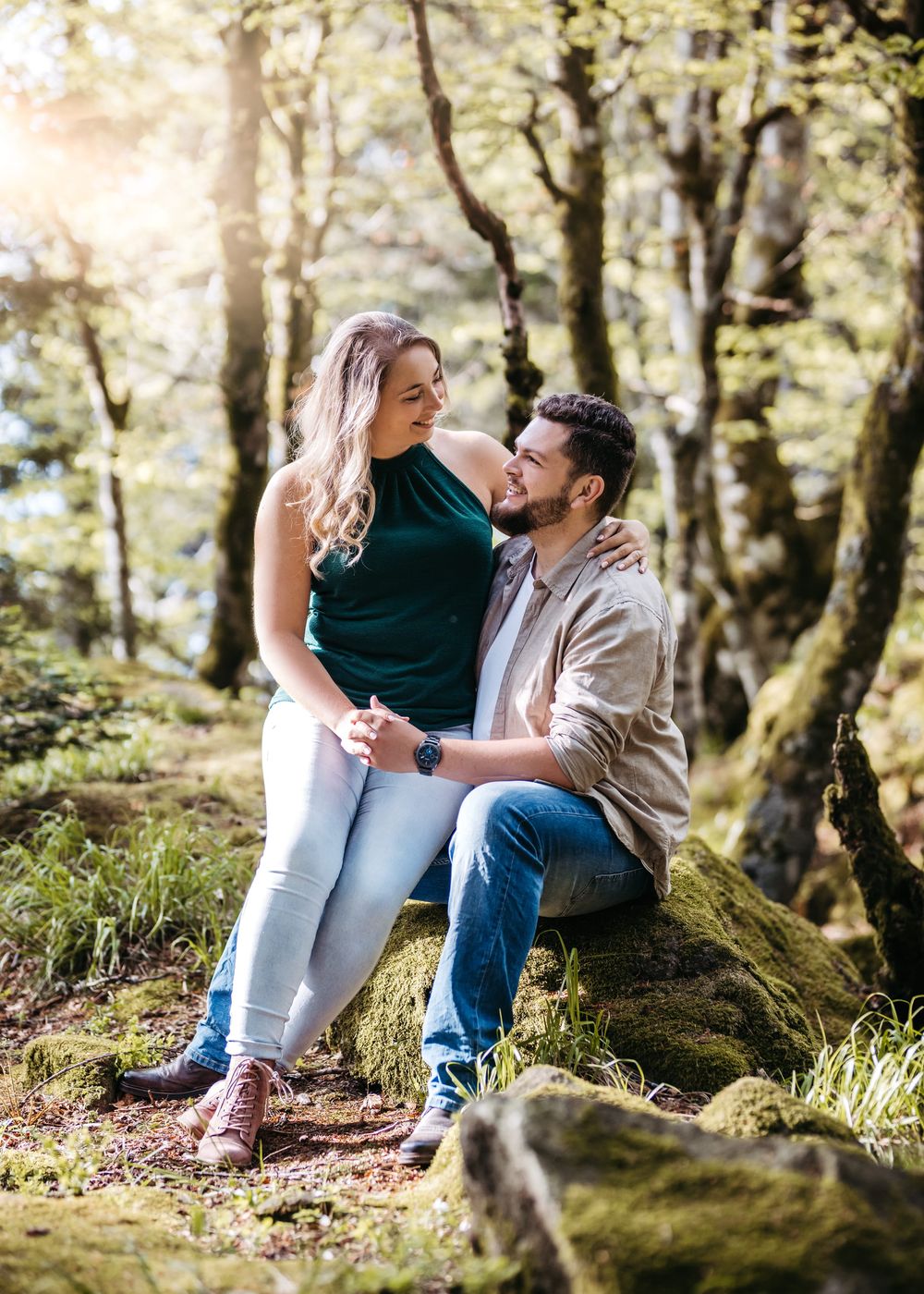A romantic couple sits on mossy rocks in a dreamy forest setting during a photography session.