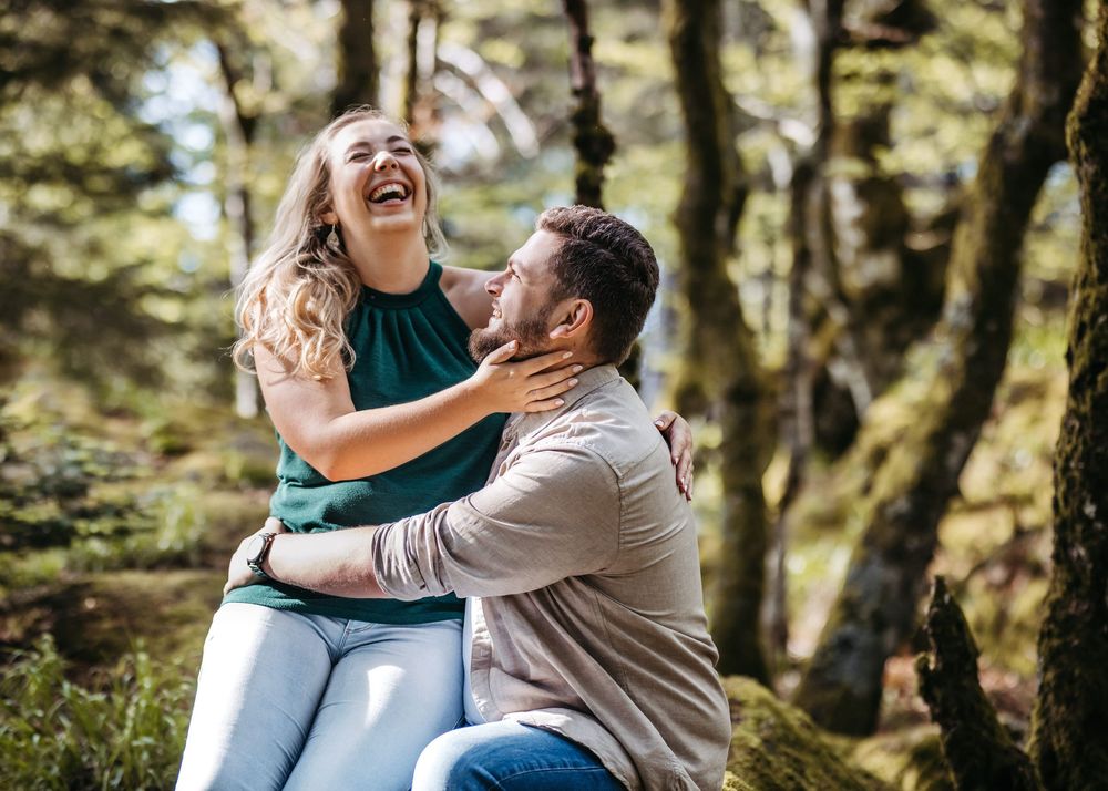 Couple laughing and embracing joyfully in a sunlit forest setting during an outdoor photo session.