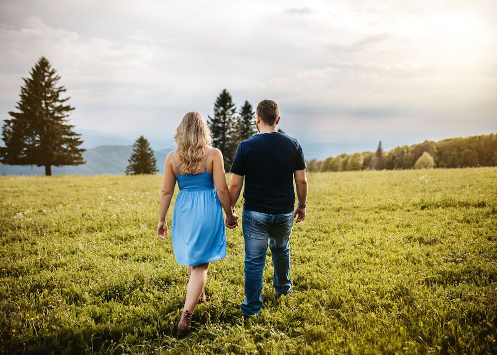 A couple walks hand in hand through a sunlit meadow at golden hour with evergreen trees in the background.
