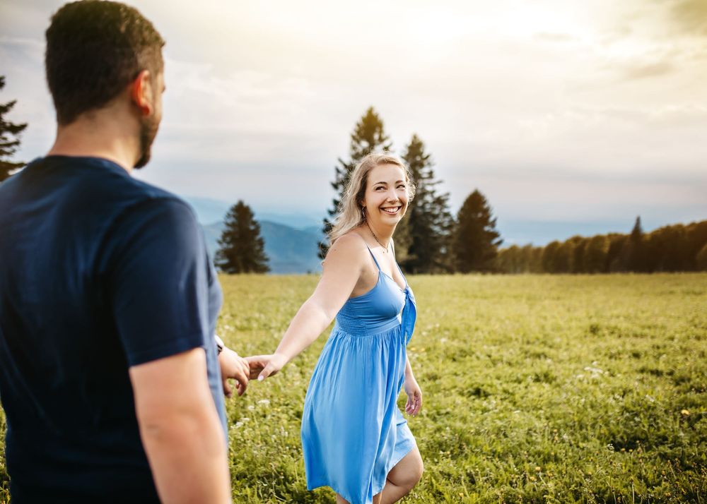 A couple holds hands while walking through a sunlit meadow with evergreen trees in the background.