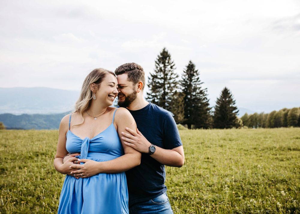 A couple in casual attire embraces while standing in a sunny meadow with evergreen trees and mountains in the background.
