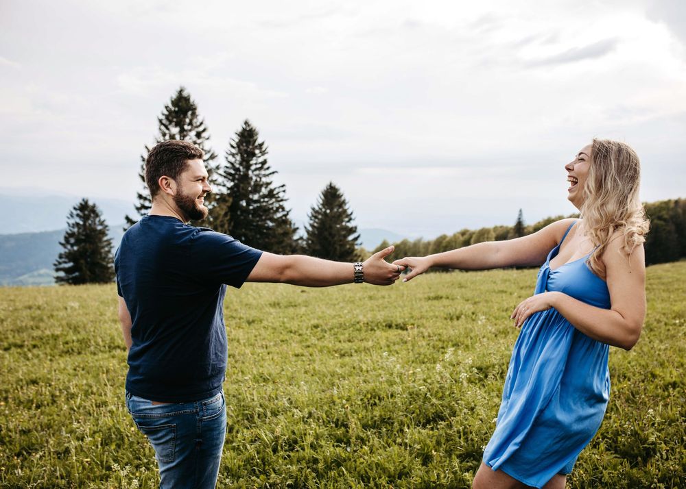 Two people hold hands and dance together in a grassy meadow with evergreen trees and mountains in the background.