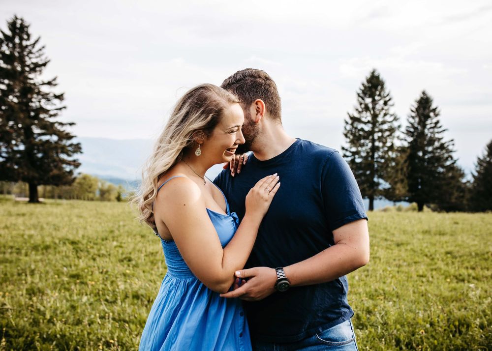 A romantic couple embraces in a grassy meadow with evergreen trees in the background on a cloudy day.
