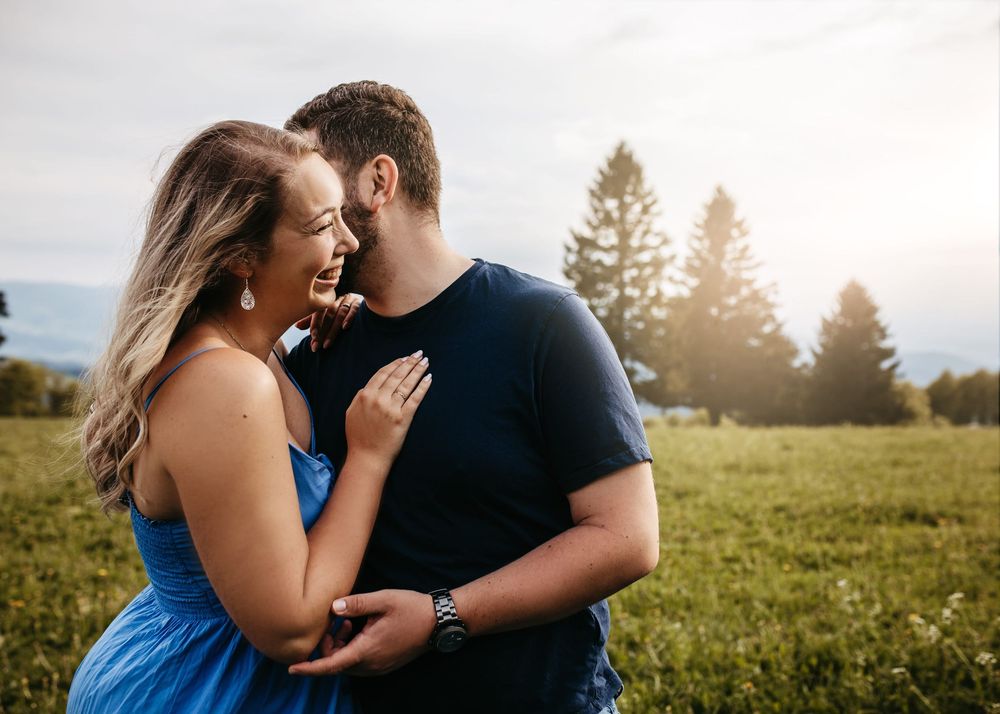 A joyful couple shares a tender moment while laughing together in a sunlit field with pine trees behind them.
