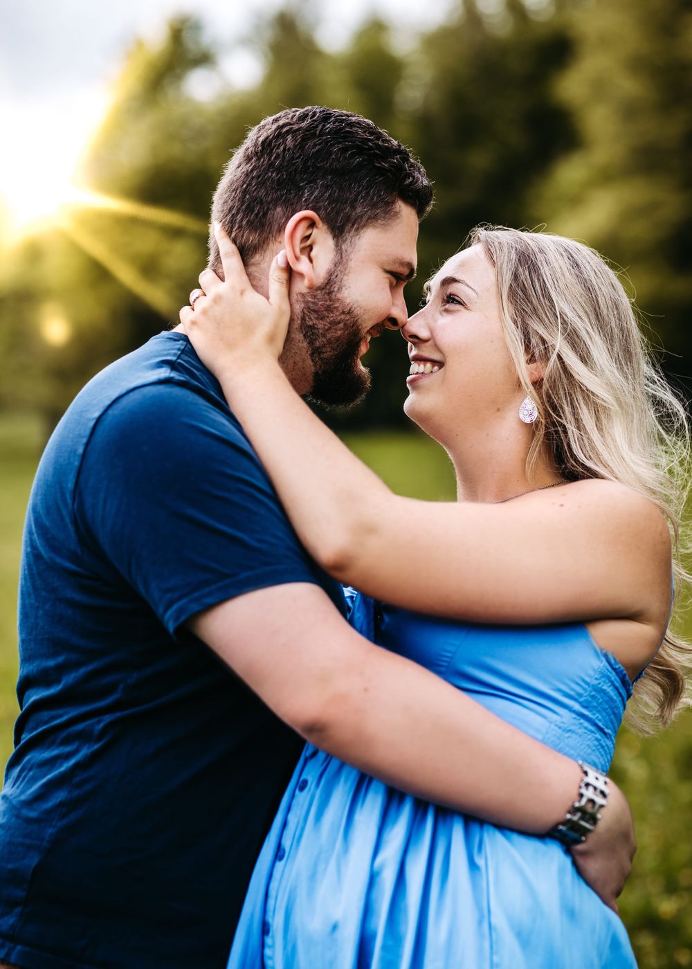 A romantic couple in blue outfits laughs and embraces while taking engagement photos outdoors in natural lighting.