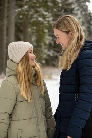 Two people wearing winter coats stand together in a snowy forest setting, sharing a candid moment.