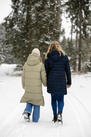 Two people in winter coats walking together on a snowy path through an evergreen forest.