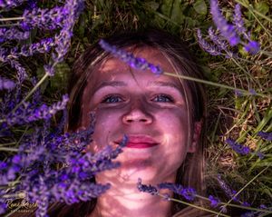 A person lies in a field of purple lavender flowers while smiling at the camera in warm sunlight.