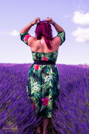 A figure in a tropical print dress stands in a vibrant purple lavender field on a sunny day.