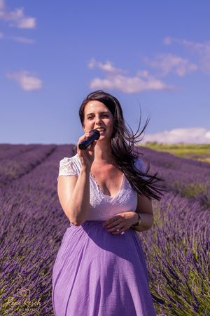 A person in a purple dress poses expressively in a blooming lavender field against a bright blue summer sky.