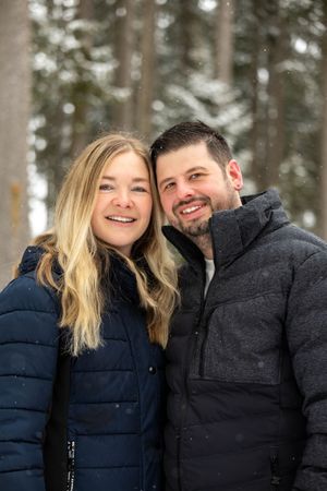 A couple in winter coats smiles and laughs together in a snowy forest setting.