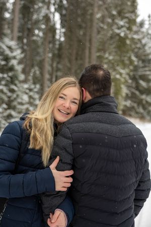 A couple in puffy winter coats share a tender moment in a snowy forest setting.