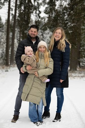 A family poses together on a snowy winter path in the forest wearing warm coats and casual attire.