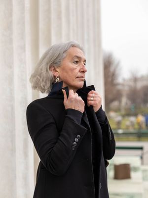 Elegant person with silver hair wearing a black coat poses thoughtfully against white columns outdoors.
