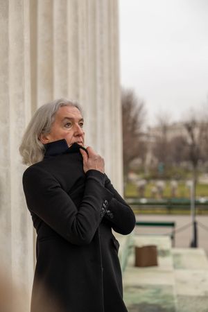 Person in black coat stands contemplatively by classical stone column on overcast day.