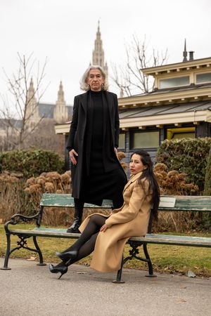 Two people in contrasting black and tan coats pose on park bench with Gothic architecture in background.