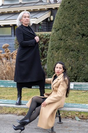 Two people in winter coats pose playfully on steps near trimmed hedges and a pool in an outdoor setting.