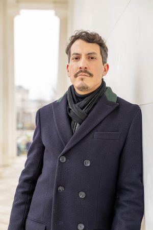 Person wearing a dark navy peacoat and scarf stands against a white architectural backdrop.