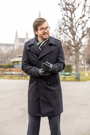 A person in a black peacoat and glasses poses outdoors in winter against a blurred background with bare trees.