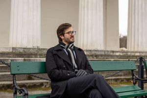 A person in a dark coat sits on a green park bench during a winter day.