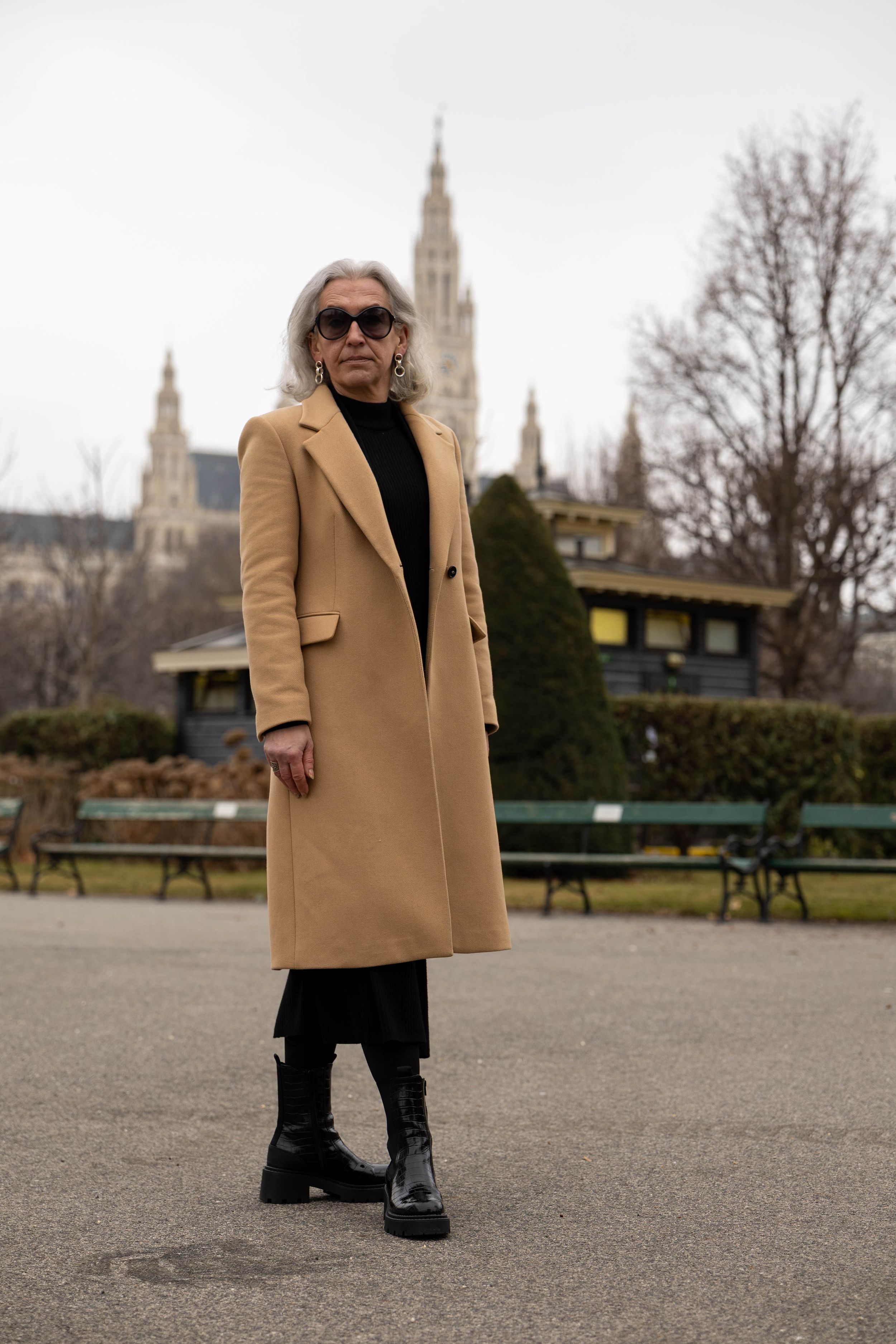 Elegant winter outfit featuring a camel coat, black boots and sunglasses with Vienna's historic architecture in the background.