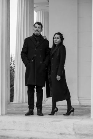A couple in black winter coats poses between white columns in a series of black and white photographs.