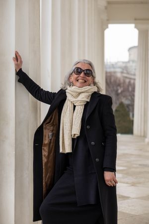 A stylish person in a black winter coat and cream scarf poses playfully between white columns on a bright day.