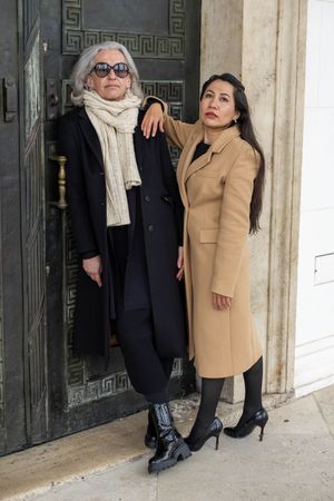 Two people pose together against a white wall wearing contrasting black and tan coats.