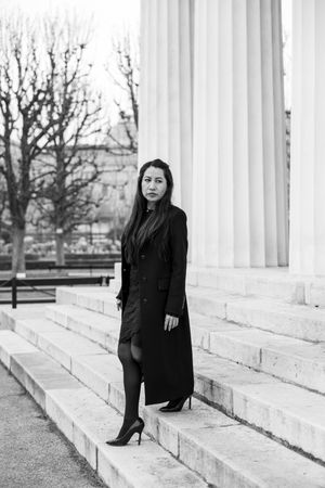 Figure in black attire poses on white marble steps with columns in a dramatic black and white composition.