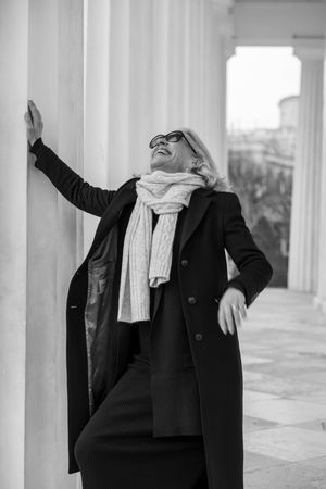 Black and white photo of person in winter coat and scarf leaning against white column while looking up and smiling.