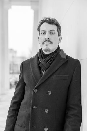 Person in black peacoat and scarf poses against white columns in black and white portrait.